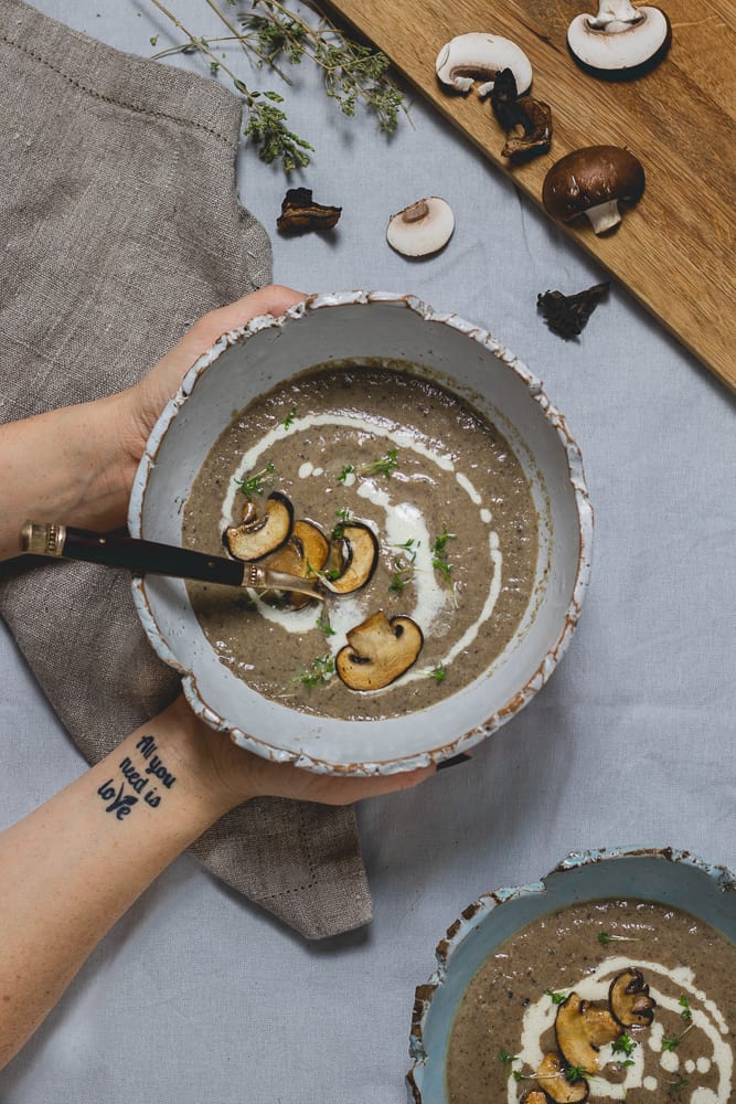 overhead shot of cream of mushroom soup in a bowl held with 2 hands, topped with vegan cream and fried mushrooms