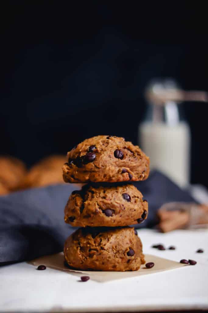 3 pumpkin chocolate chip cookies stacked, with a bottle of oat milk, spices, and more cookies on a cake rack in the background